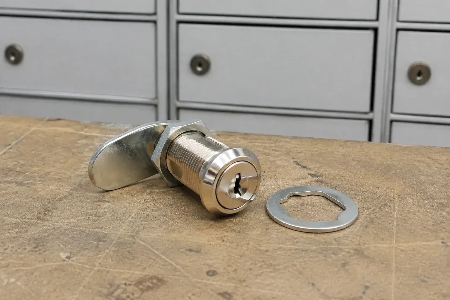 Detached threaded cam lock and mounting washer on a workbench in front of closed mail compartments. — Mailbox Lock Replacement, Welland