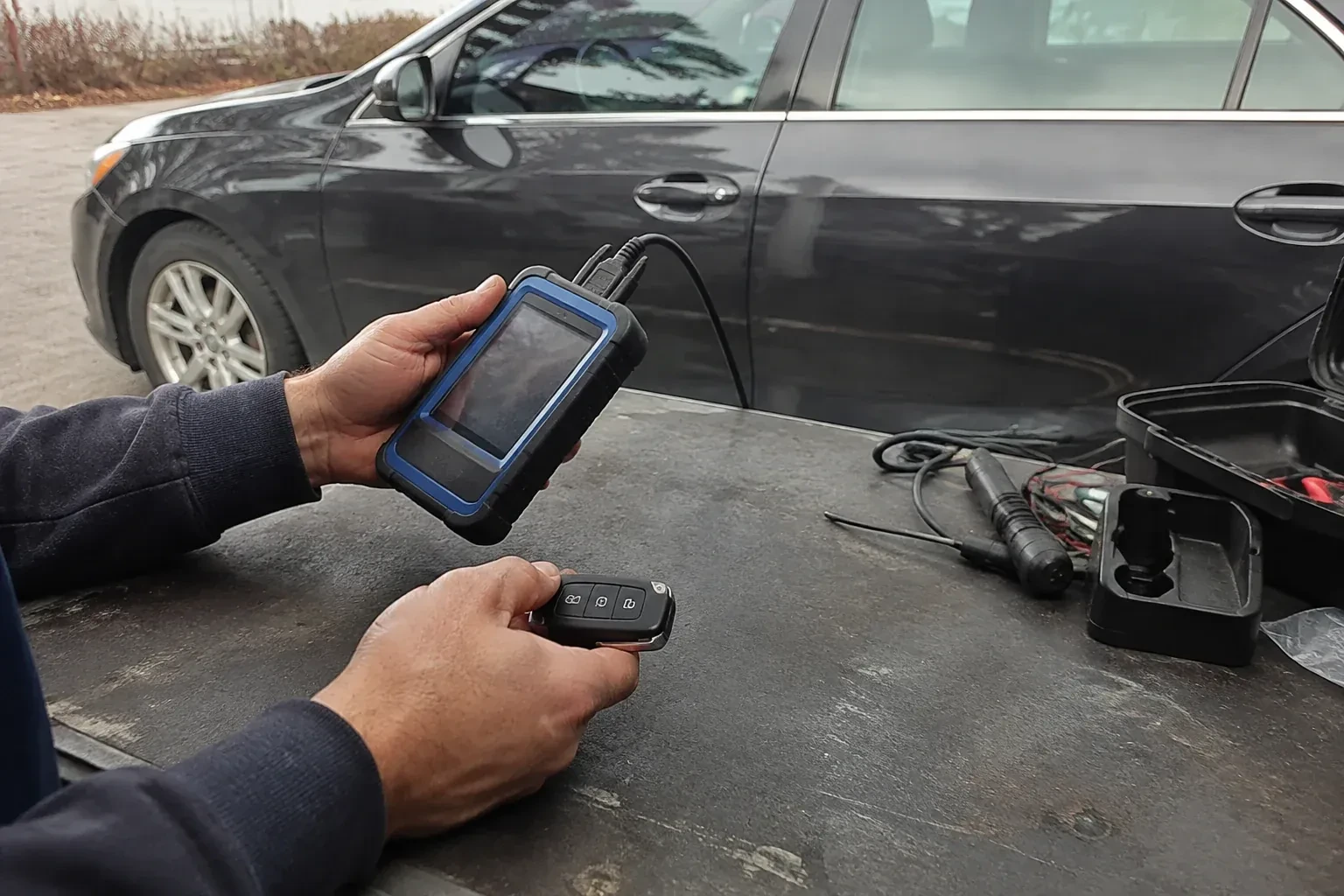 Hands holding a blue-edged electronic device and car-key fob at a worktable beside a parked car. — Transponder Key Programming, Welland